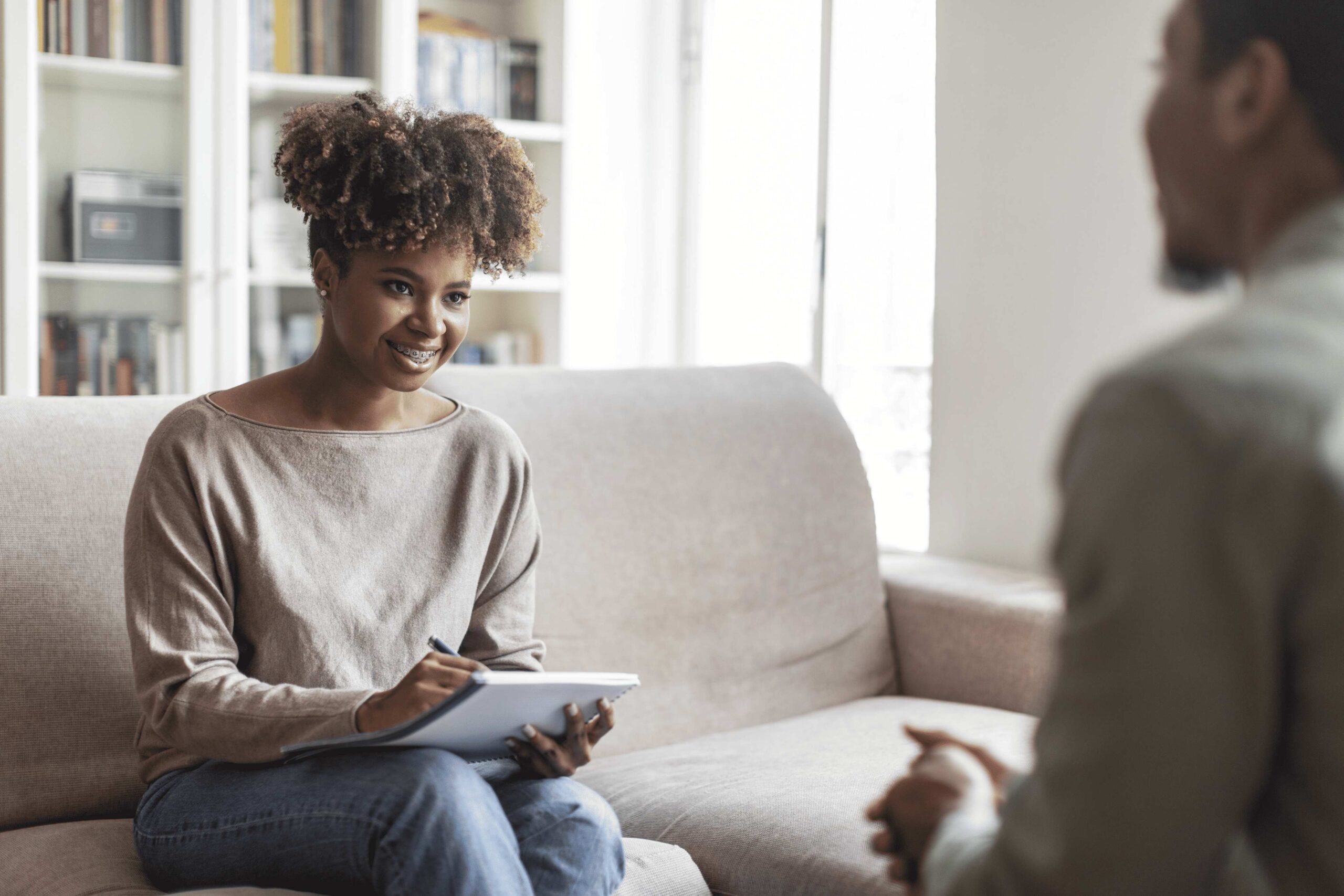 Therapist smiling while taking notes on a notepad, seated across from a client during a counseling session.