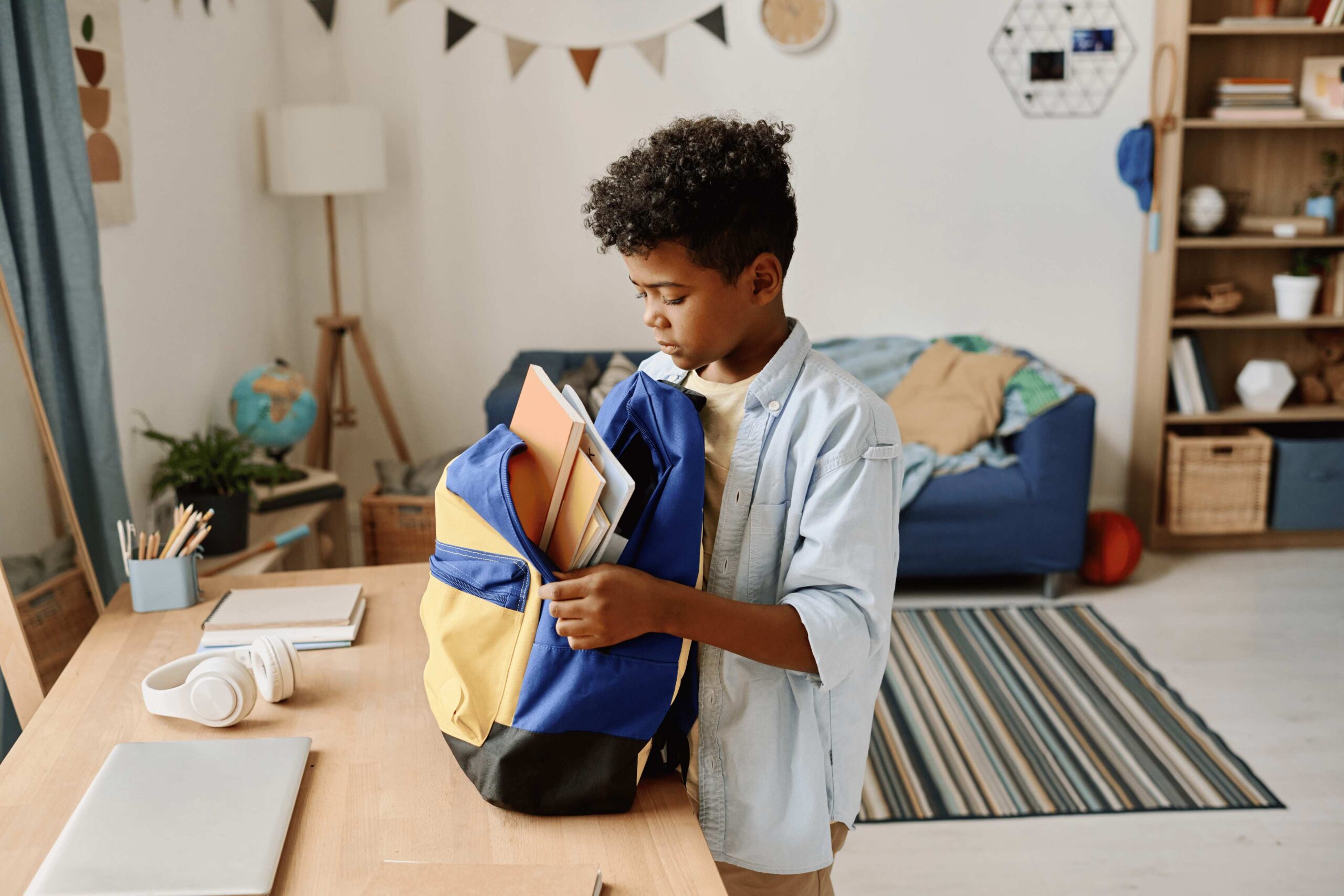 Child placing books into a backpack at a desk in a bedroom with a couch and shelves in the background.