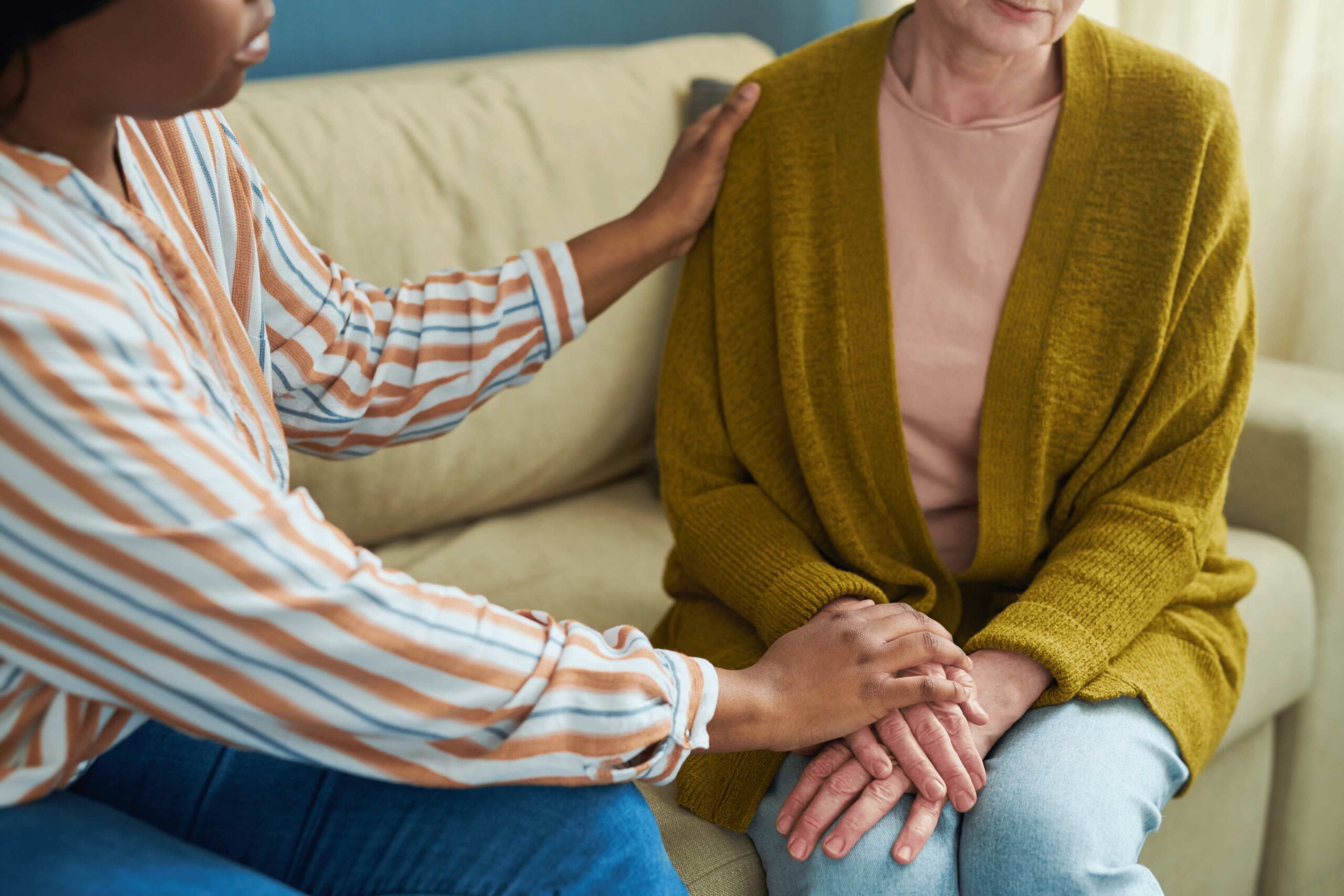 One person gently holding another’s hands and placing a supportive hand on their shoulder while sitting together on a couch.