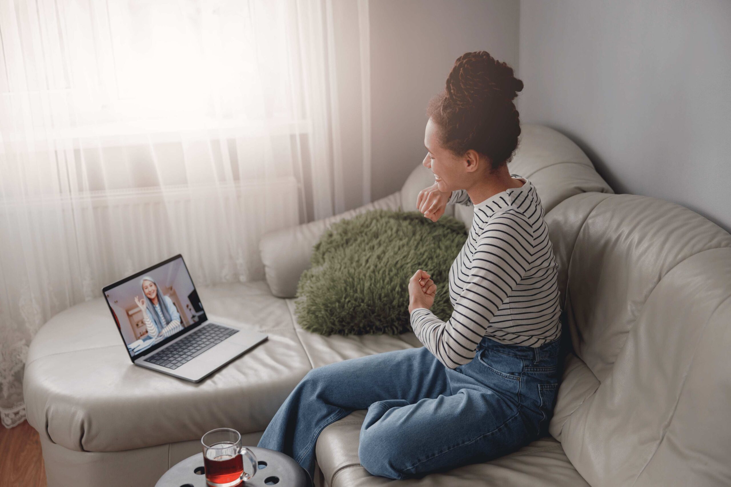 Woman sitting on a couch participating in a video call on a laptop, with a glass of tea on a nearby table.