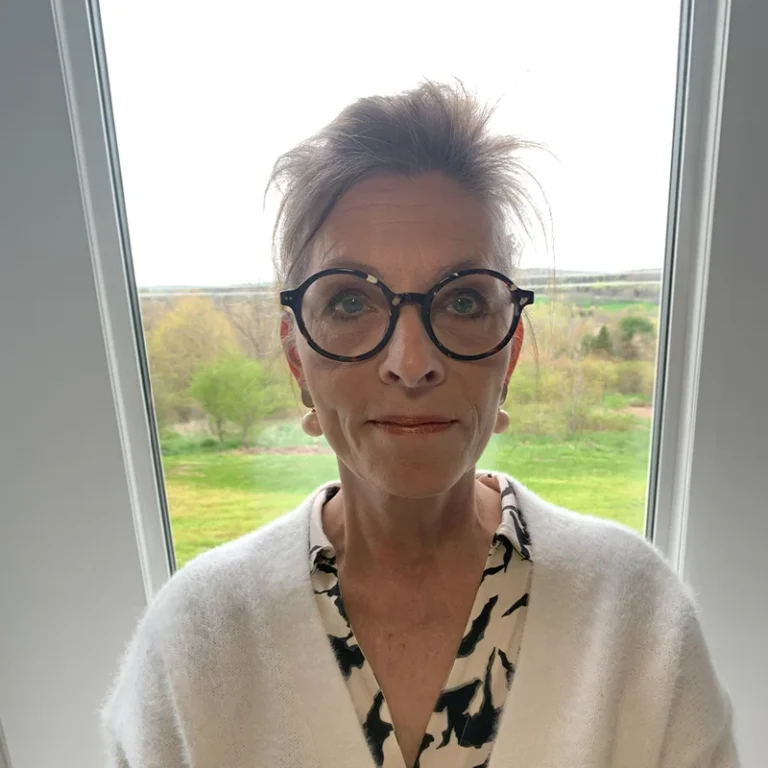 Person with glasses wearing a white cardigan, standing indoors near a window.