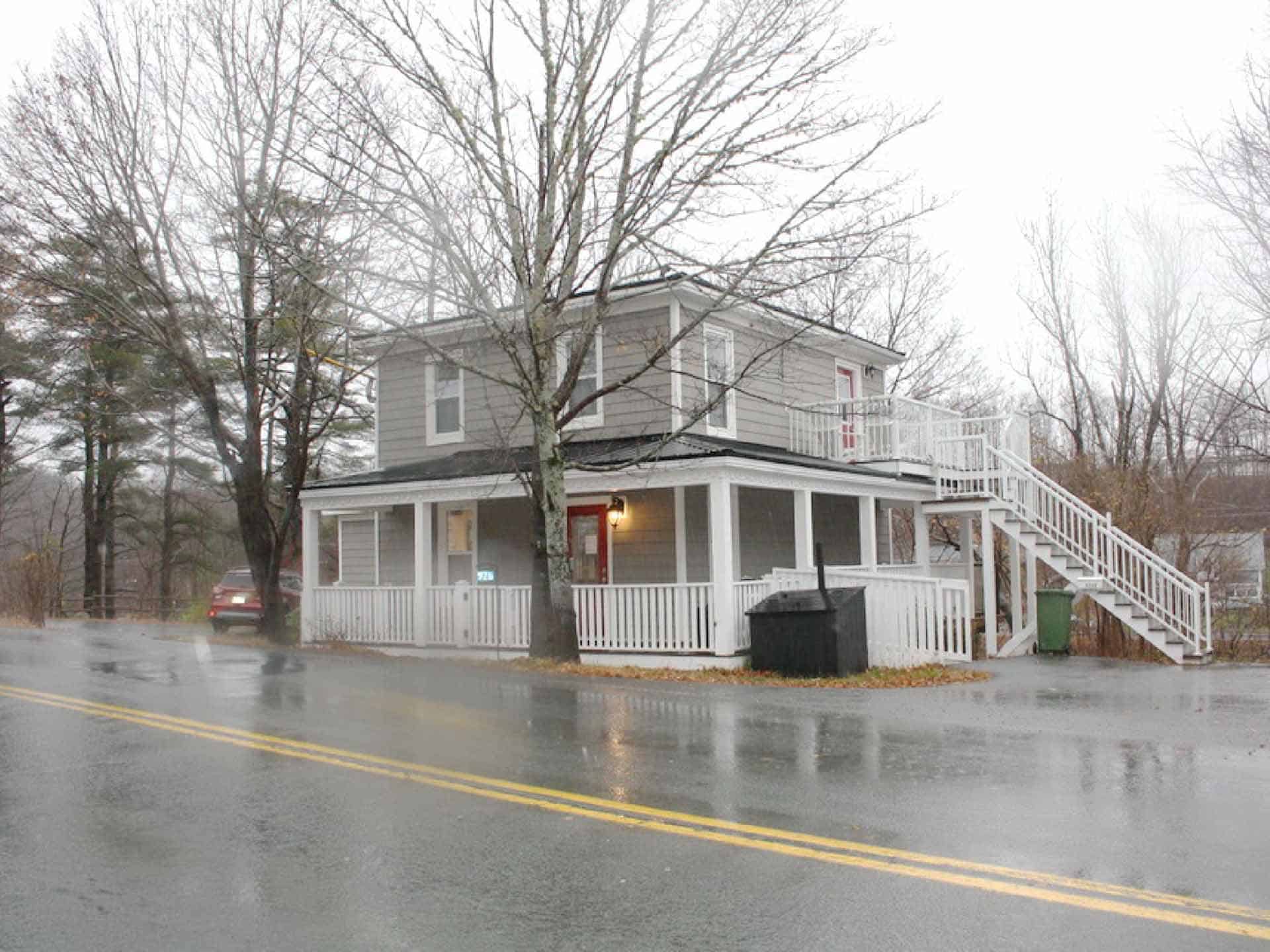Two-story building with wraparound porch beside a wet road on a rainy day.