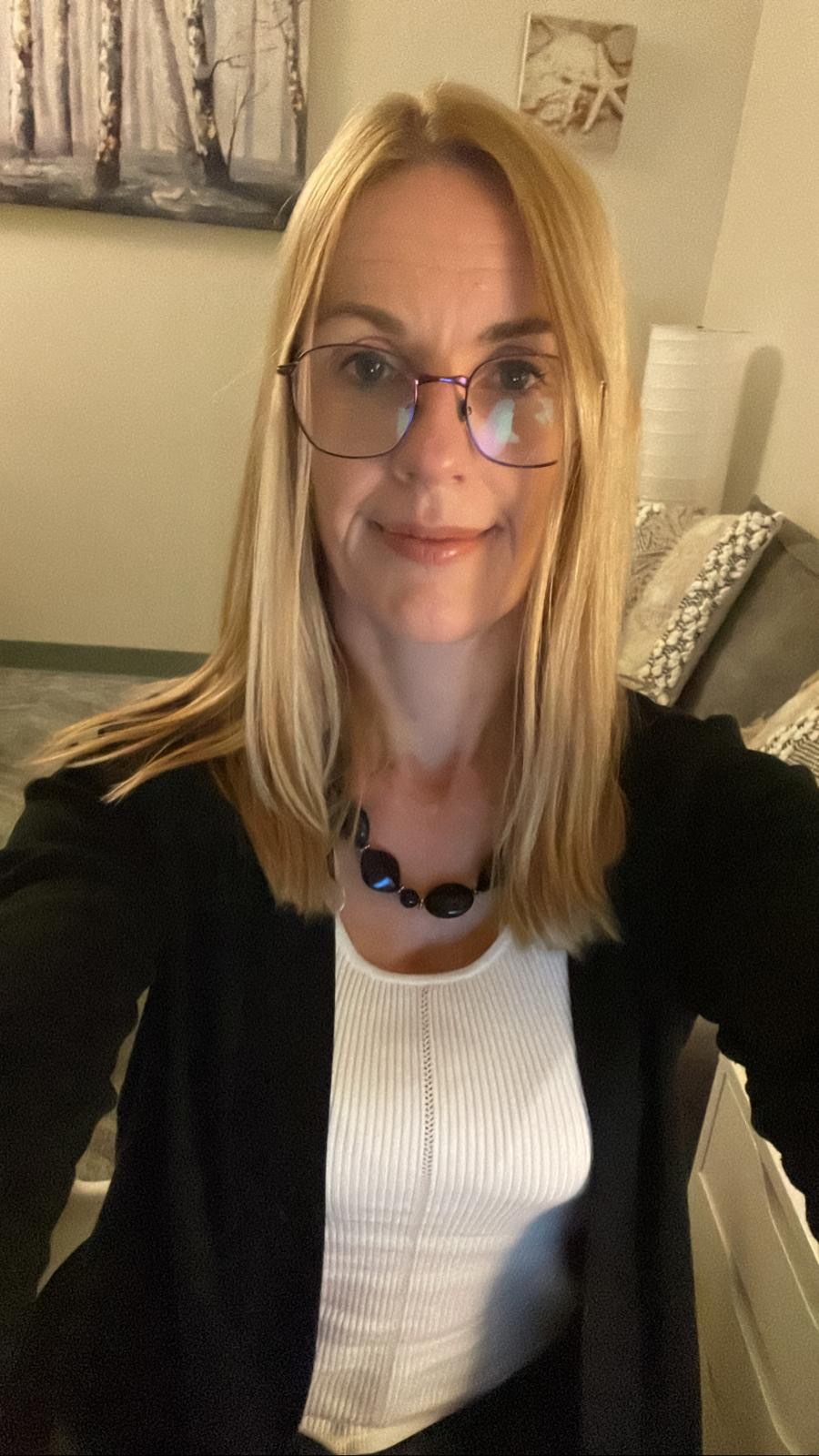 Person with blonde hair and glasses wearing a black cardigan over a white top in an indoor selfie.