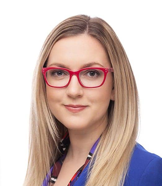 Person with red glasses and long blonde hair wearing a blue blazer against a white background.