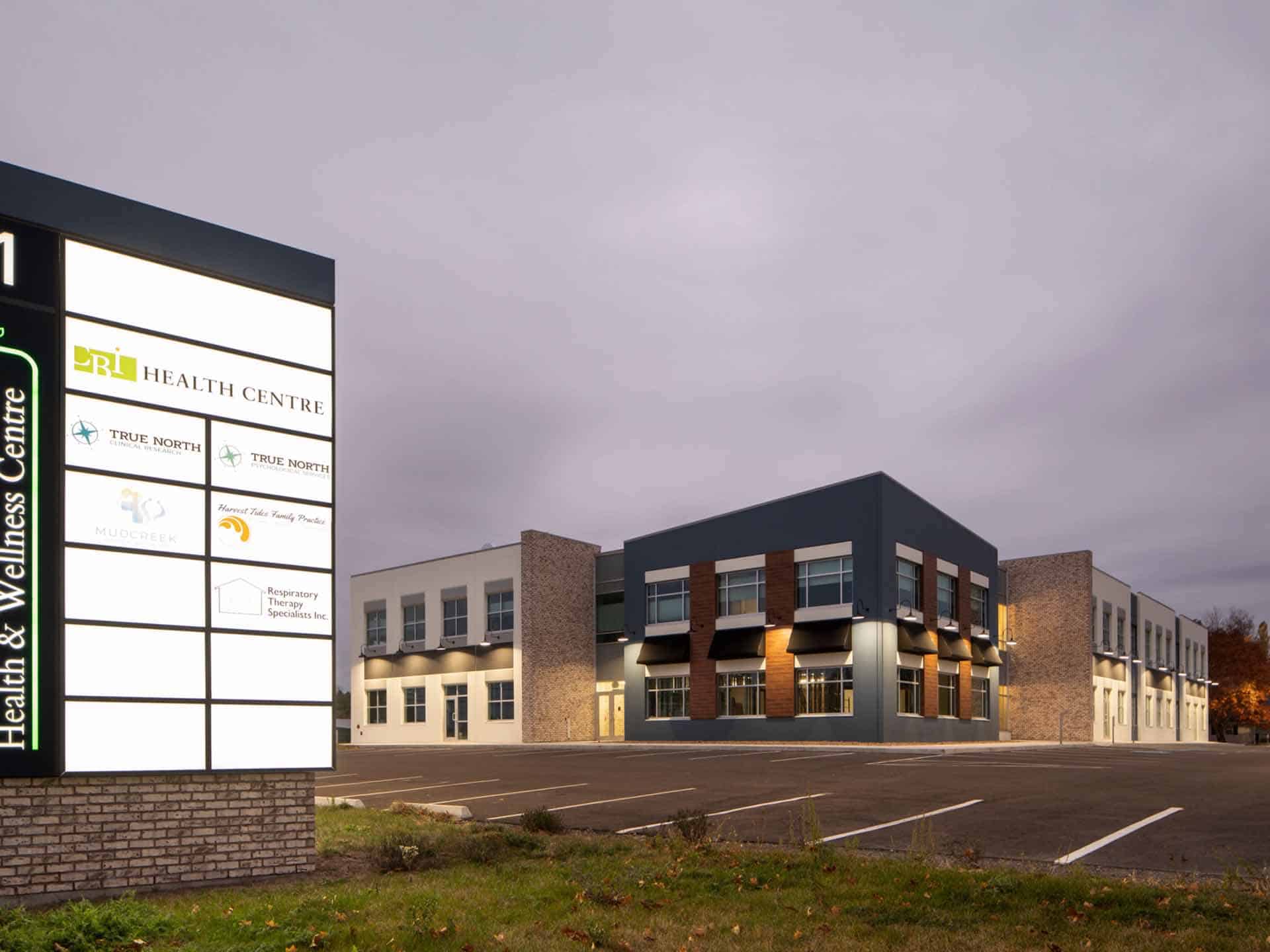 Exterior of a health and wellness centre building with a directory sign in the foreground at dusk.