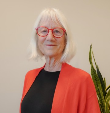 Person wearing glasses and a cardigan, standing indoors near a plant.