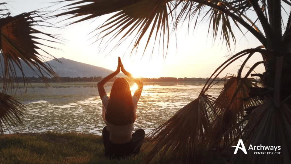 Person sitting by the water at sunrise with hands together overhead, framed by palm leaves.
