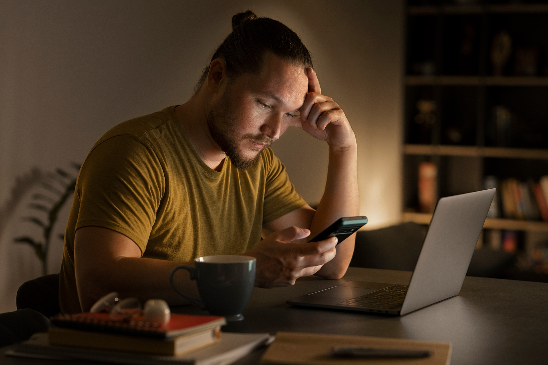 Person sitting at a desk at night, looking at a phone beside an open laptop and mug.