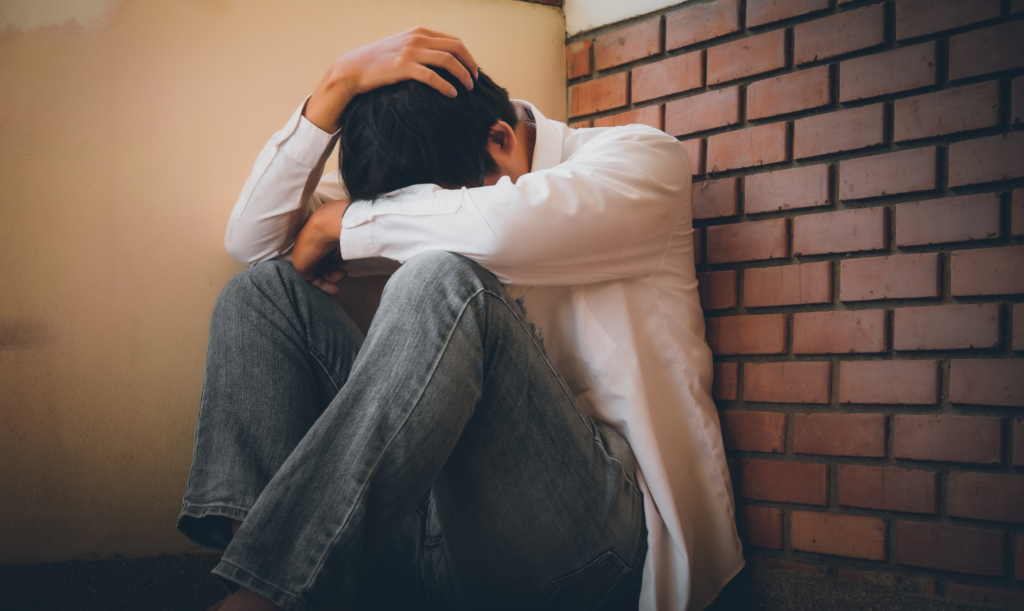 Person sitting on the floor against a brick wall with head lowered and one hand on their head.