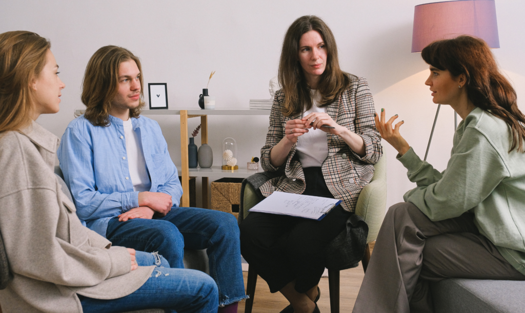Four people seated in a circle in a room, talking while one person holds a clipboard.