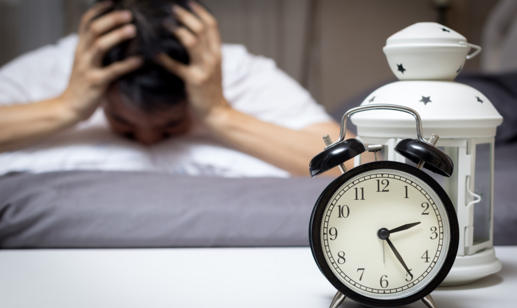 Alarm clock on a bedside surface with a blurred person in bed holding their head in the background.