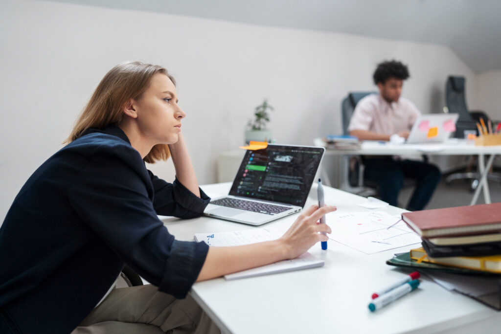 Person at a desk holding a pen and resting their head on one hand, with another person working in the background.