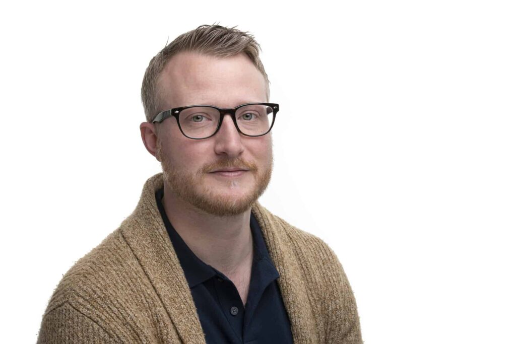 Man with glasses wearing a tan cardigan over a dark shirt against a white background.
