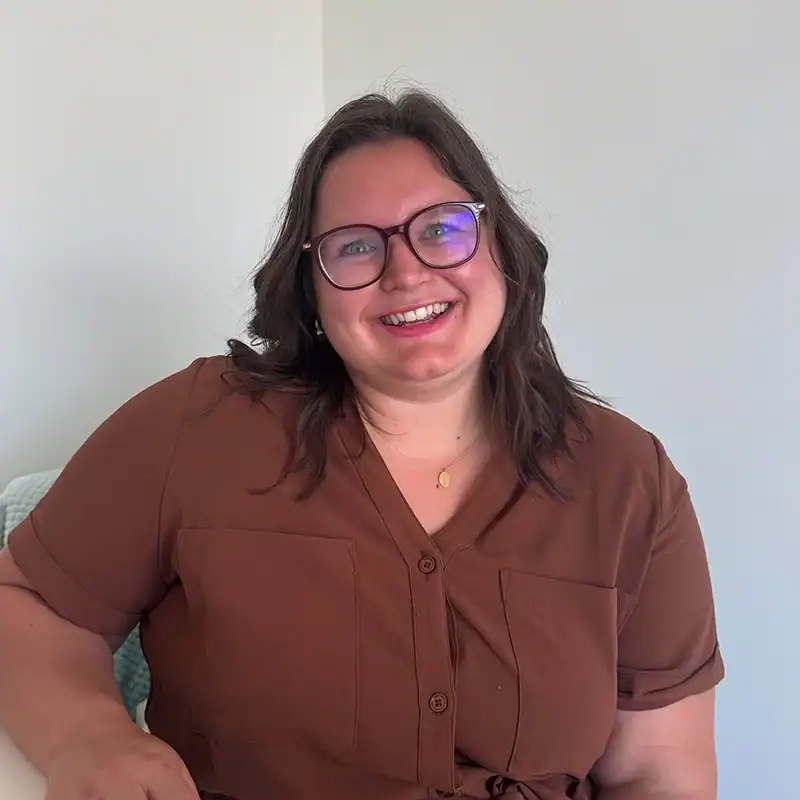 Smiling person with glasses wearing a brown button-up top against a light indoor background.