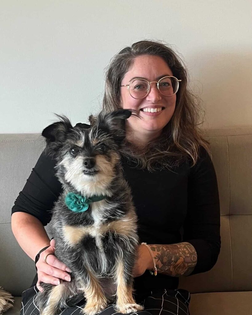 Smiling woman with glasses holding a small black-and-white dog while seated indoors.