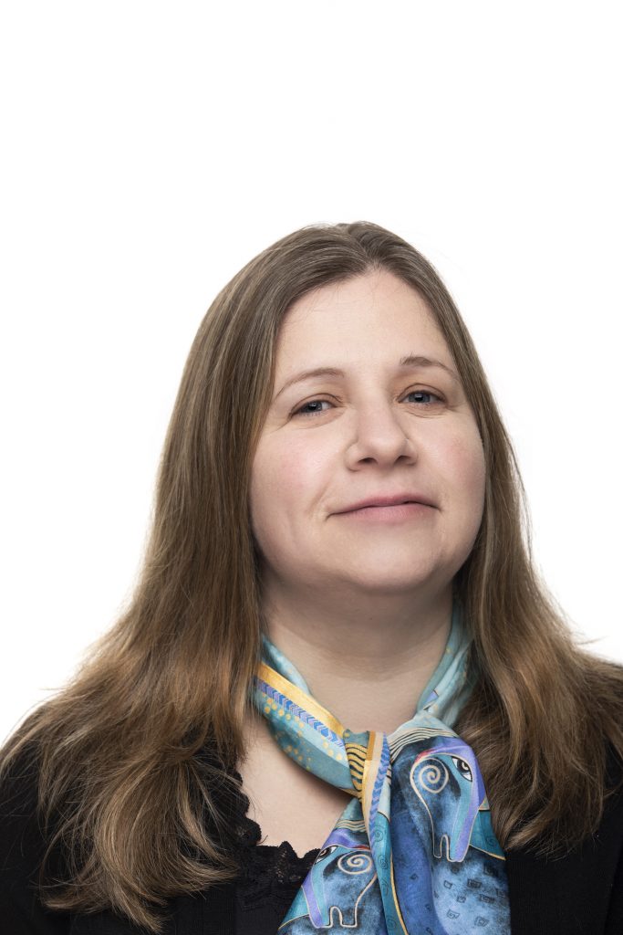 Person with long hair wearing a black top and patterned blue scarf against a white background.