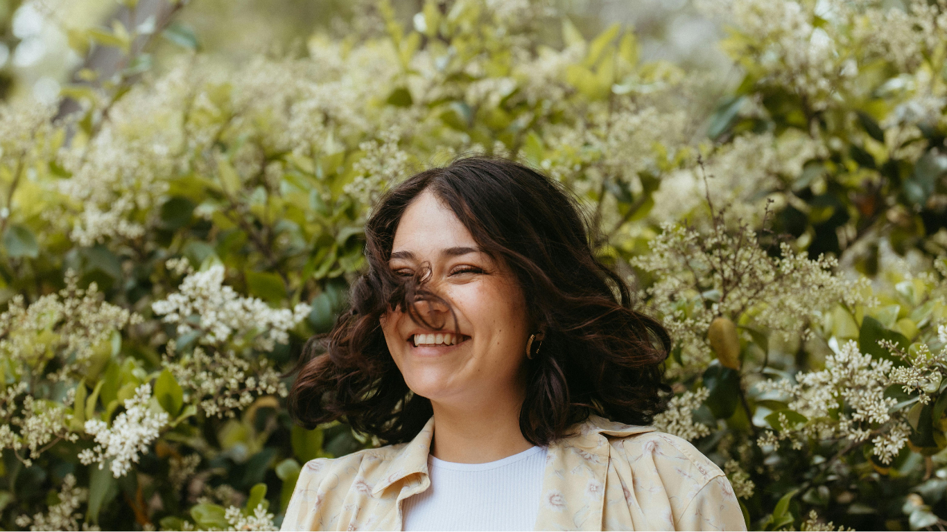 Smiling person outdoors surrounded by white flowers and green foliage.