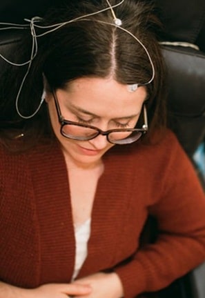 Woman wearing glasses with monitoring electrodes attached to her head, looking down while seated in a chair.